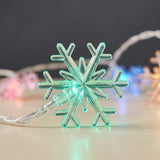 Snowflake string lights feature a translucent snowflake glowing green on a pale surface, with blurred multicolor lights against a dark background.