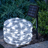 A coil of white solar string lights on a stone surface next to a black solar panel stake, with green garden bushes in the background.