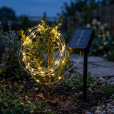 Warm white solar string lights loop around a small garden shrub beside a black solar panel on a stake at dusk.