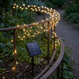Warm white solar string lights draped over a curved metal garden railing, with a small solar panel stake visible in the garden bed at dusk.
