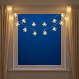 Warm white snowflake-shaped string lights draped across a window with beige curtains on both sides, set against a deep blue sky background.