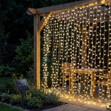 Warm white LED curtain string lights hanging from a wooden pergola in a backyard patio setting, with a small solar panel stake visible on the left.