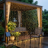 Warm white LED curtain string lights hanging in a wooden patio gazebo, illuminating a wet deck and outdoor furniture at dusk.
