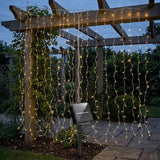 Solar-powered LED curtain string lights draped over a wooden garden pergola at dusk, with a small solar panel stake visible on a stone patio in the background.