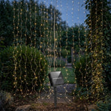 Warm white solar curtain light hangs across a leafy garden path, with a black solar panel on a ground stake.