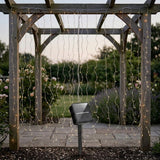Warm white solar curtain light hangs from a wooden pergola above garden paving, with a black solar panel in front.