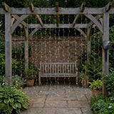 Warm white solar curtain light hangs beneath a wooden pergola before a wooden bench, with its solar panel beside the right post.