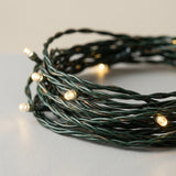 Coiled Christmas string lights with a dark green wire and warm white glowing bulbs, photographed on a light gray surface.