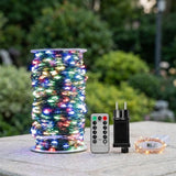 A multicolor LED string light on green cable is wound around a spool beside a remote, black adapter, and small coil outdoors.