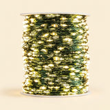 A spool of green wire Christmas string lights glowing with warm yellow lights, set against a solid light beige background.
