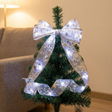 A glowing silver ribbon light string forms a large bow and loose loops around a small Christmas tree beside a sofa.