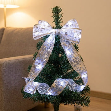 A glowing silver ribbon light string forms a large bow and loose loops around a small Christmas tree beside a sofa.