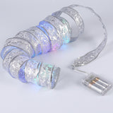 Silver ribbon light string glows in multiple colors, arranged in loose coils beside a clear battery box on a light gray background.