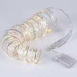 A silver patterned ribbon light string with warm white lights curls beside a clear battery pack on a pale gray background.