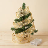 A gold ribbon light string wraps around a small Christmas tree, with its connected clear battery box resting on the wooden tabletop.
