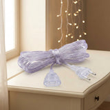 A transparent extension cord rests bundled on a cream table, with both connectors visible and warm string lights behind.