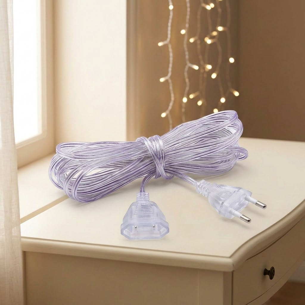 A transparent extension cord rests bundled on a cream table, with both connectors visible and warm string lights behind.