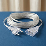 A coiled clear plastic extension cord with transparent plugs, resting on a blue surface with a beige curtain in the background.