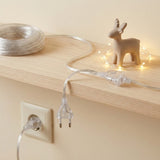 A transparent extension cord lies partly coiled on a wood shelf beside a deer ornament and string lights, with its plug hanging.