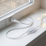 A transparent extension cord loops across a white windowsill, connecting an outlet to warm glowing string lights.