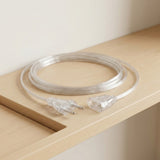 A transparent extension cord rests loosely coiled on a pale wood corner shelf, with its two-round-pin plug and female connector visible.
