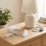White star-shaped fairy string lights with a clear battery pack and power plug, styled on a light wood bedside table next to a cream-colored table lamp.