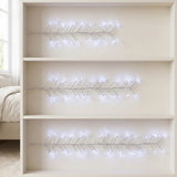 Three sets of white LED branch-shaped string lights displayed on a beige three-shelf bookcase, with a bed and window visible in the background.