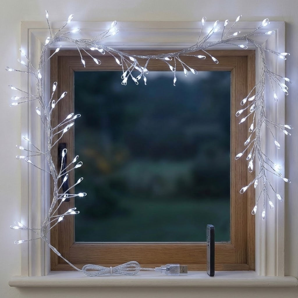 White LED fairy string lights shaped like bare branches draped around a wooden window frame, with the lights turned on and a coiled cable and small black remote on the windowsill.