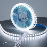 A coiled white LED light strip with a blue backing, shown on a dark reflective surface with its attached power cable and switch.