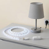 A white LED strip light coiled on a white table next to a gray table lamp, with a white USB controller and power cord nearby. The background is a plain beige wall.