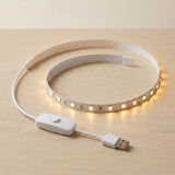 A flexible white LED light strip coiled on a light wood surface, illuminated with a warm yellow glow. The strip includes a white USB connector and a white inline switch module.