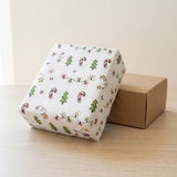 White wrapping paper with green trees, candy canes, stars, and string lights covers a gift leaning against a brown box on wood.