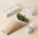 White Christmas wrapping paper appears as stacked sheets, a wrapped box, bouquet lining, and a twine-tied parcel on a pale wooden tabletop.