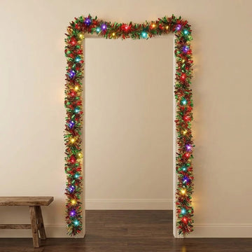 A rectangular doorway decorated with a green artificial holiday garland wrapped in warm white and multicolored Christmas lights, shown against a beige wall with a wooden floor and a small bench at the lower left.