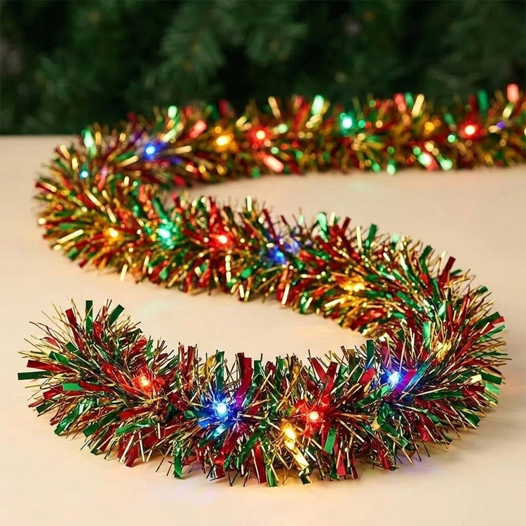 A festive Christmas tinsel garland with colorful lit bulbs, arranged in a U-shape on a light beige surface with a blurred green holiday background.