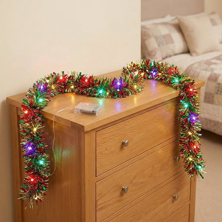 A heart-shaped multicolor tinsel Christmas garland with built-in LED lights draped over a wooden dresser in a bedroom setting.
