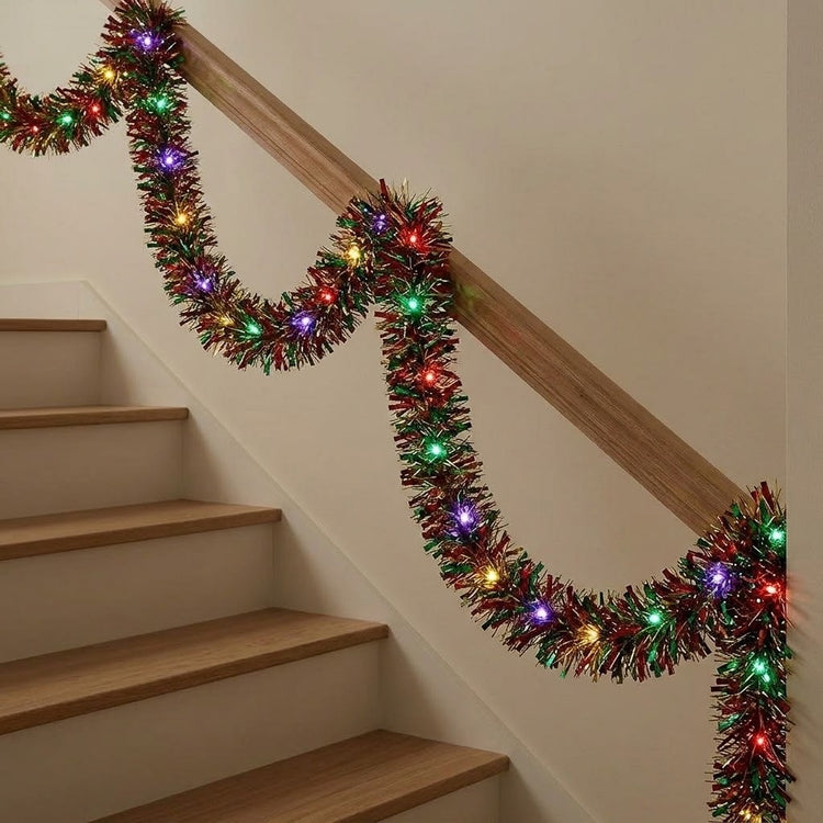 A festive Christmas tinsel garland with multicolored lights draped over a wooden staircase railing in an indoor setting.
