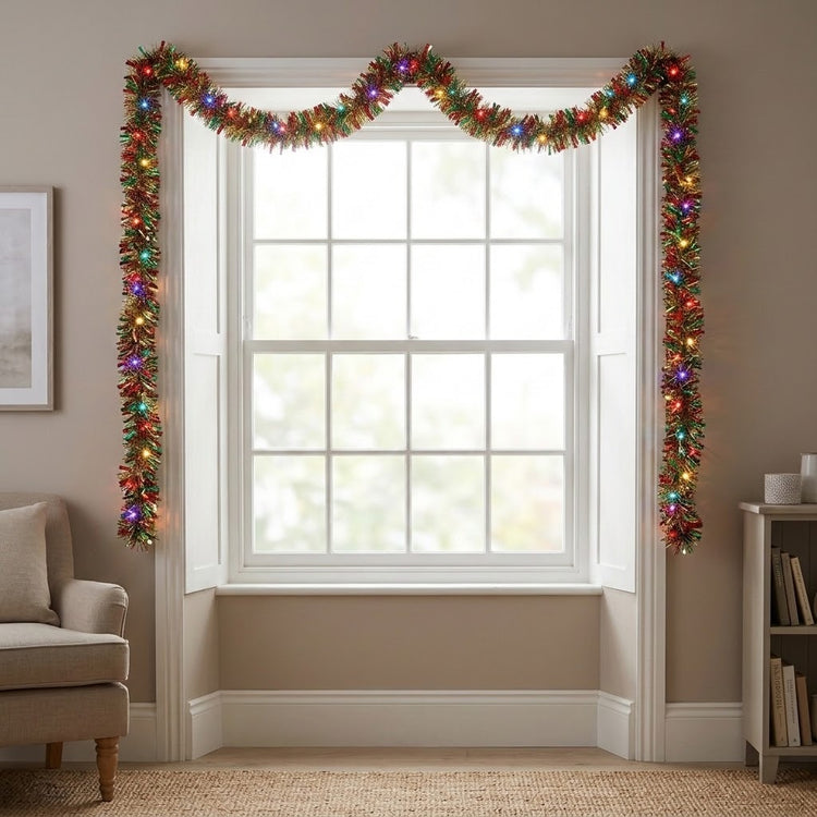 A festive Christmas window garland with a scalloped valance and vertical panels, decorated with multicolored lights and green tinsel, shown hanging on a white window in a cozy living room setting.