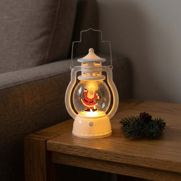A white decorative lantern with illuminated dancing Santa rests on a wooden side table beside evergreen sprigs and pinecones.