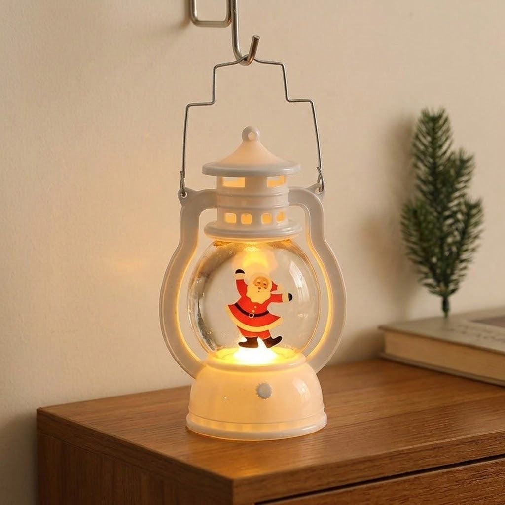 A white decorative lantern with dancing Santa hangs from a hook above a wooden cabinet beside a book and evergreen sprig.