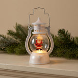 A white decorative lantern with dancing Santa and warm light sits on a pale wooden mantel in front of evergreen branches.