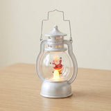 A silver decorative lantern with a clear globe and illuminated Santa stands on a wooden tabletop against a plain cream wall.