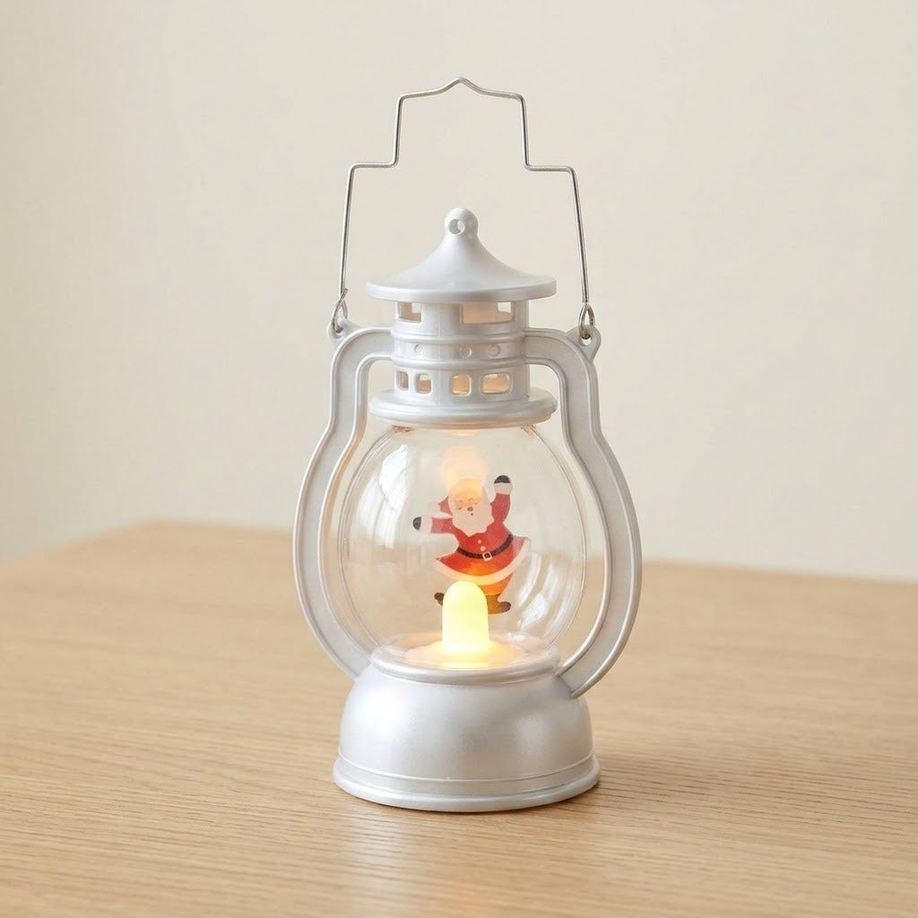 A silver decorative lantern with a clear globe and illuminated Santa stands on a wooden tabletop against a plain cream wall.
