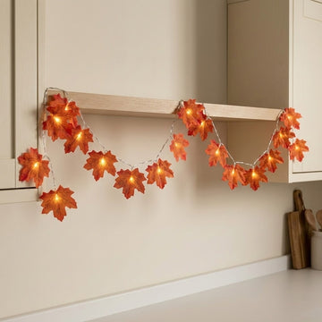 Autumn leaf string lights with warm glowing bulbs draped over a wooden shelf in a kitchen setting.