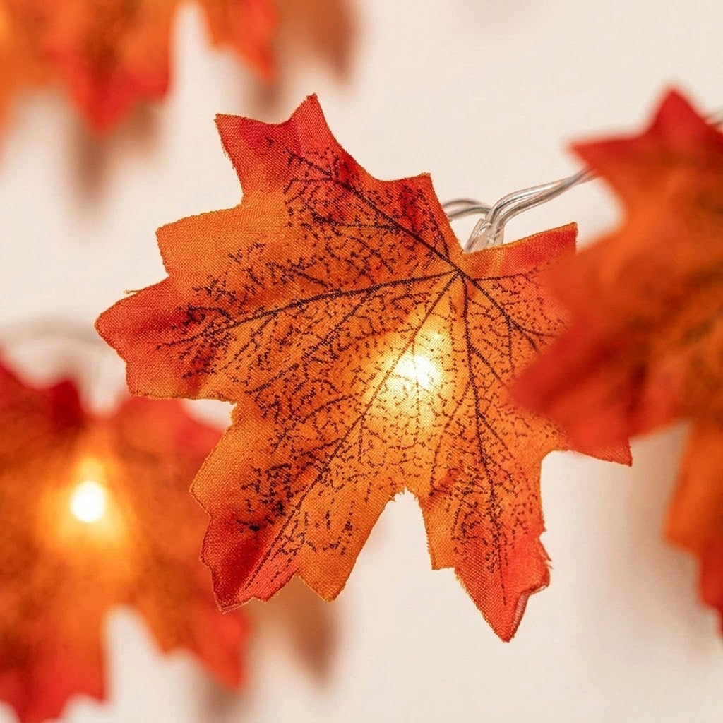 Warm white LED string lights shaped like autumn maple leaves, glowing against a soft off-white background.