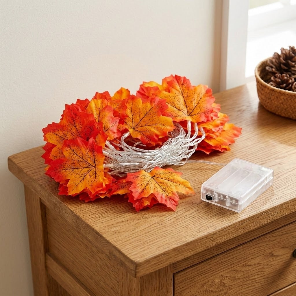 Maple leaf string lights with orange, red, and yellow leaves sit coiled beside a clear battery box on a wooden table.