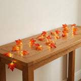 Warm white string fairy lights shaped like orange and red autumn leaves draped across a wooden console table against a plain off-white wall.