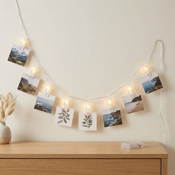 Warm white photo clip lights hold landscape photographs and botanical cards above a wooden cabinet, with a clear battery box resting below.
