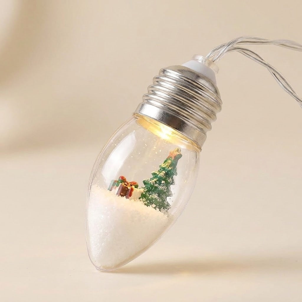 Christmas string lights feature a clear, silver-capped bulb containing a miniature green tree and gift, shown close up against a cream background.