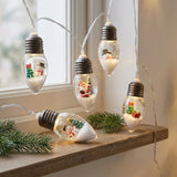 Christmas string lights with clear bulbs containing festive figurines hang beside a window above a wooden shelf with evergreen sprigs.